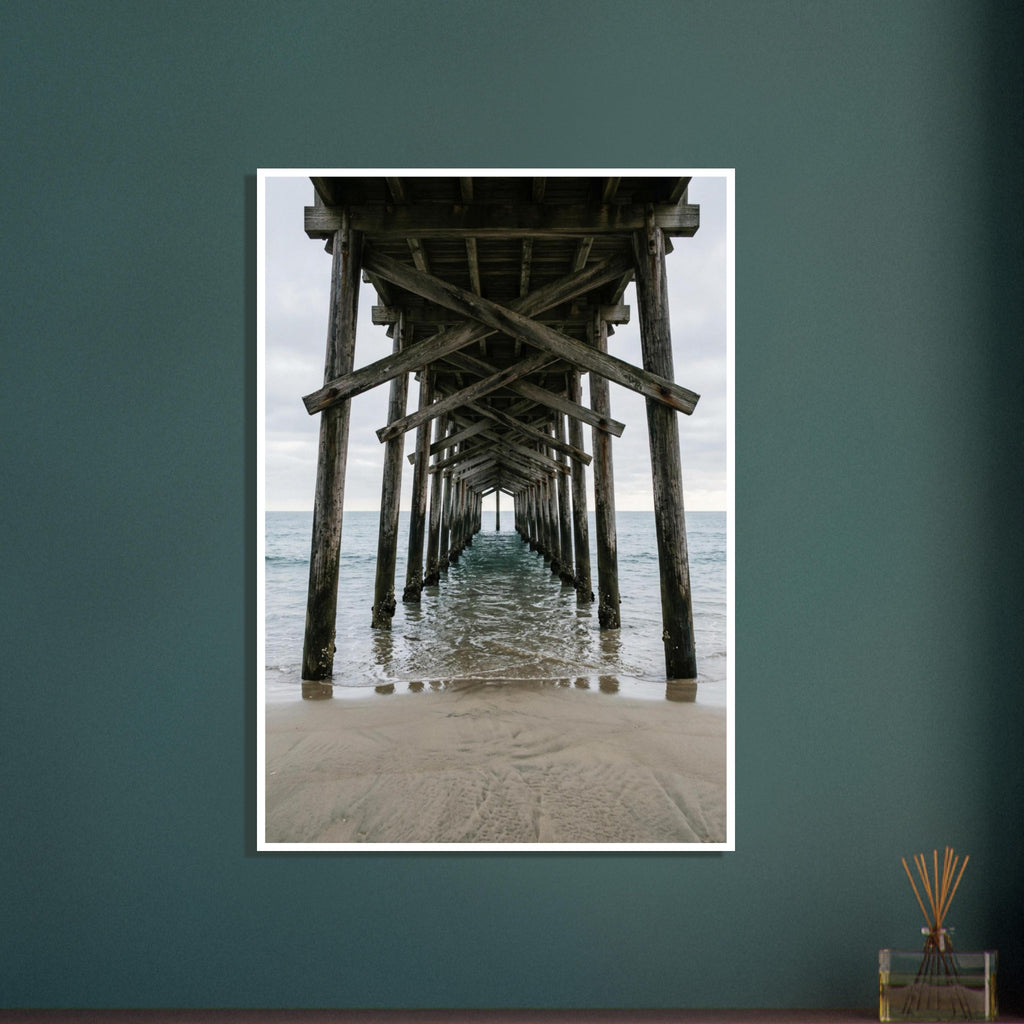 Timber pier viewed from beneath, with crossed wooden beams receding over gentle sea waves and wet sand under a grey sky.