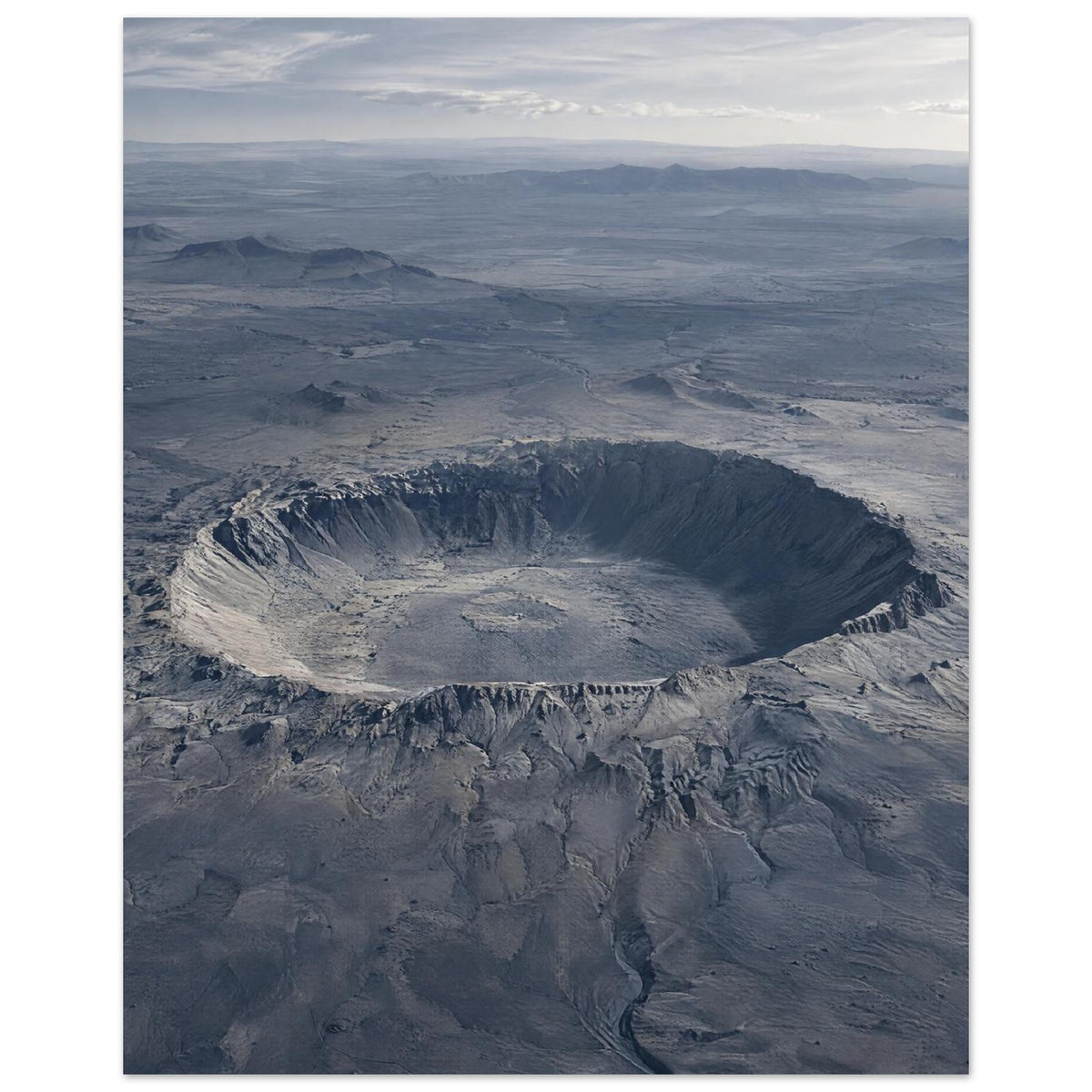 Large circular crater in a blue-grey desert plain, with rugged slopes, distant hills and a cloudy sky.