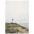 Lighthouse on a rocky headland beyond grassy sand dunes, with gentle waves and a wide, pale grey sky.