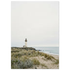 Lighthouse on a rocky headland beyond grassy sand dunes, with gentle waves and a wide, pale grey sky.