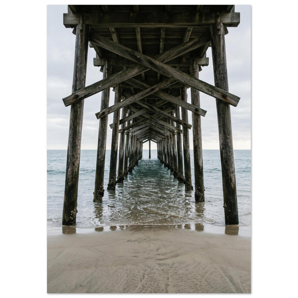 Timber pier viewed from beneath, with crossed wooden beams receding over gentle sea waves and wet sand under a grey sky.