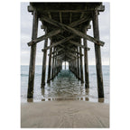 Timber pier viewed from beneath, with crossed wooden beams receding over gentle sea waves and wet sand under a grey sky.