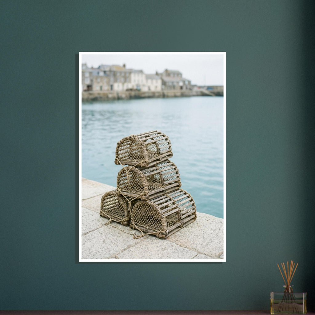 Four wooden lobster pots stacked on a stone quay beside pale blue harbour water, with blurred waterfront buildings beyond.