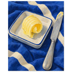 The Butter Curl still life showing a curled pat of butter on a rectangular dish beside a silver knife on cobalt cloth.