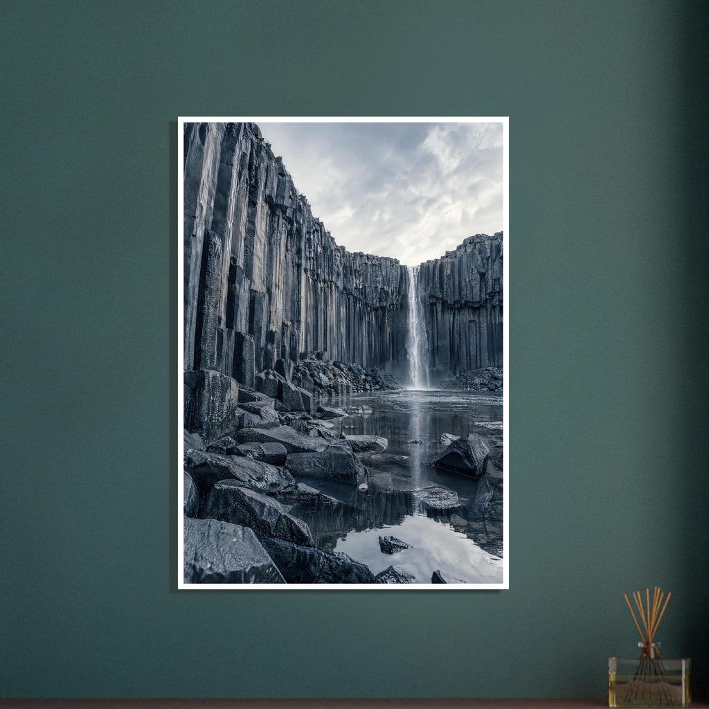 Waterfall cascading between tall basalt columns into a reflective pool, with dark foreground rocks and a cloudy sky.