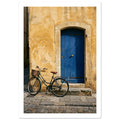 Green bicycle with a woven basket beside a blue wooden door in a golden plaster wall, above worn stone paving.