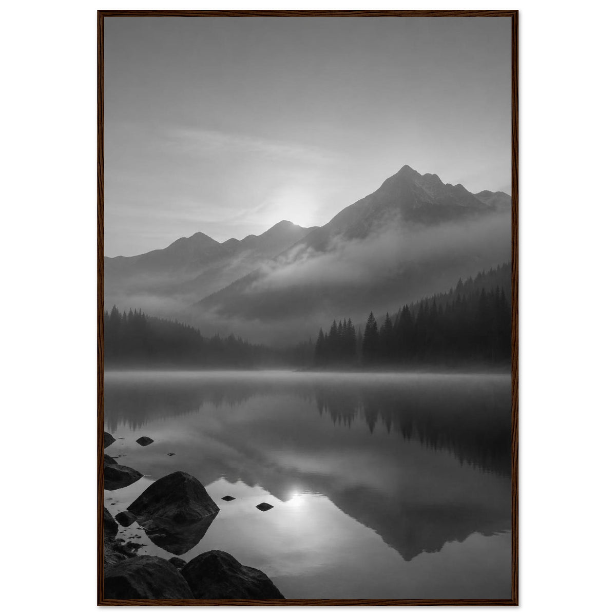 Mountain peaks and conifer forests reflected in a misty lake, with rocks in the foreground, in black and white.