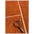 Tennis racket and yellow-green ball on an orange clay court, with white lines, footprints and strong shadows.