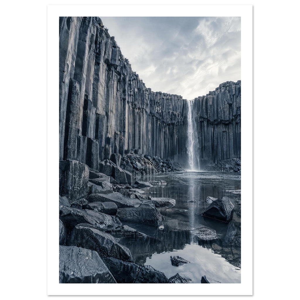 Waterfall cascading between tall basalt columns into a reflective pool, with dark foreground rocks and a cloudy sky.