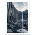 Waterfall cascading between tall basalt columns into a reflective pool, with dark foreground rocks and a cloudy sky.