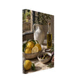 Lemons, olives, figs and a bottle of oil on a stone table beside a cream jug of leafy branches and a sunlit window.