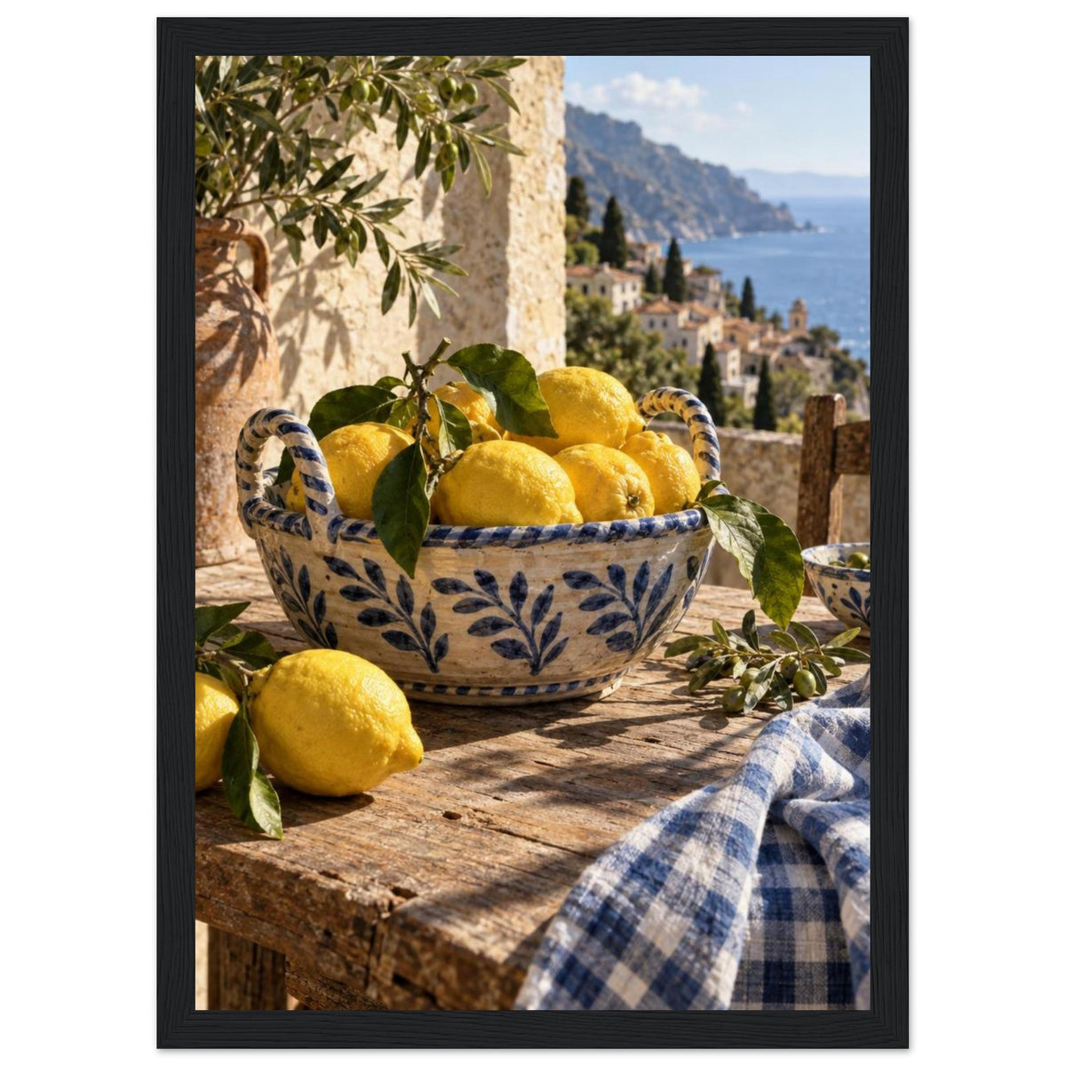 Lemons in a blue-and-white bowl on a wooden table, with olive branches, checked cloth and a coastal village beyond.