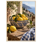 Lemons in a blue-and-white bowl on a wooden table, with olive branches, checked cloth and a coastal village beyond.