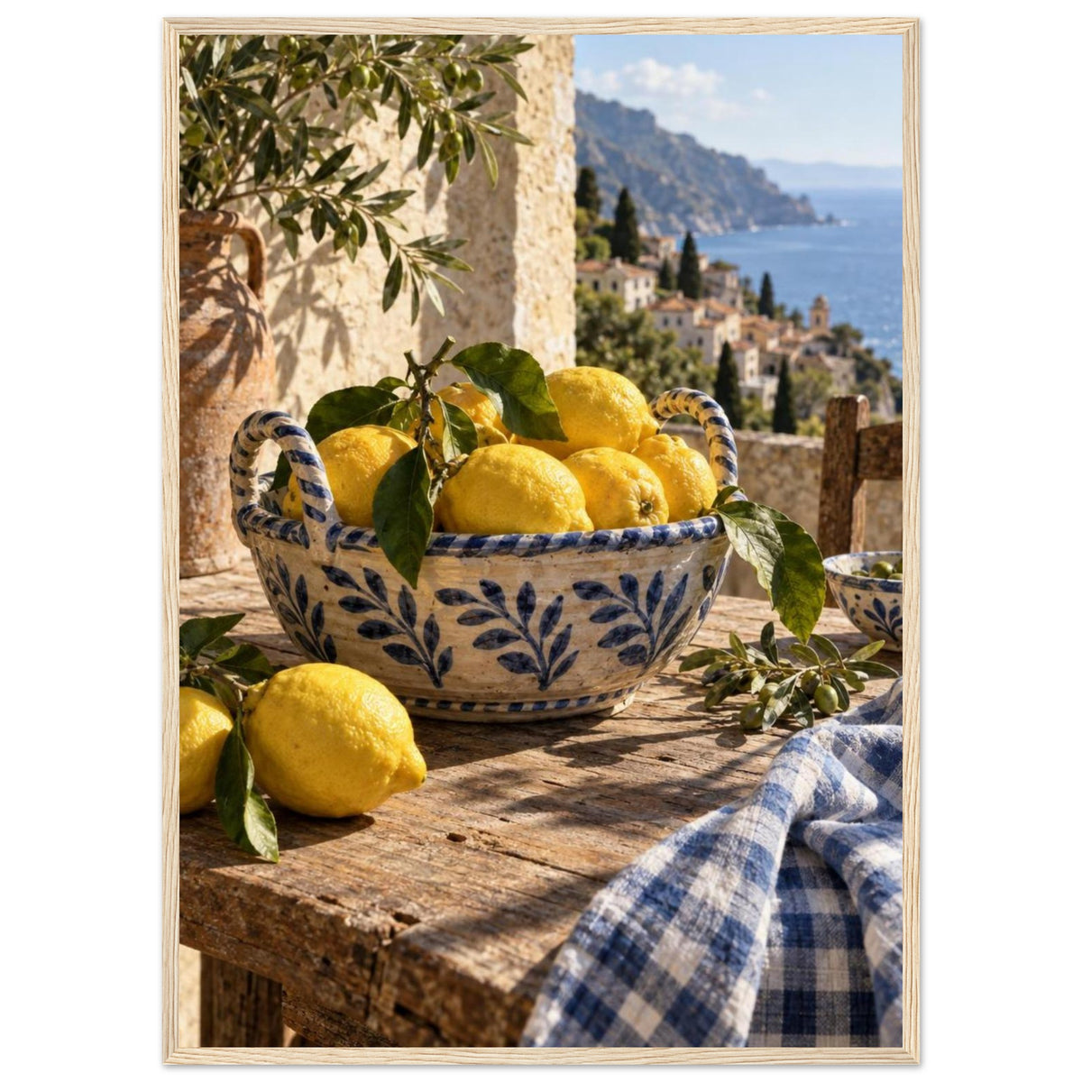 Lemons in a blue-and-white bowl on a wooden table, with olive branches, checked cloth and a coastal village beyond.