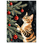 Striped orange cat reaching into evergreen branches with red Christmas baubles against a dark background with snowflakes.