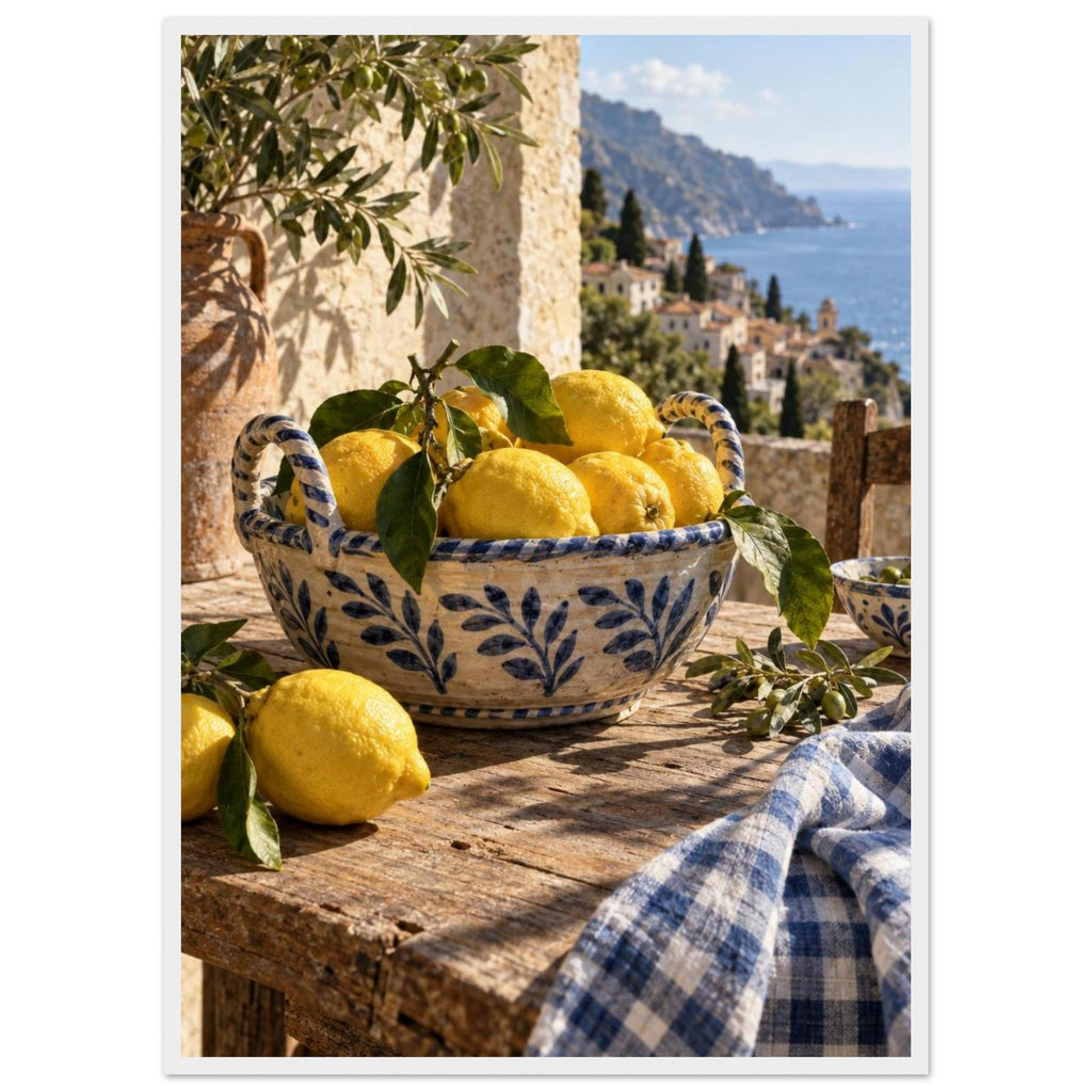 Lemons in a blue-and-white bowl on a wooden table, with olive branches, checked cloth and a coastal village beyond.