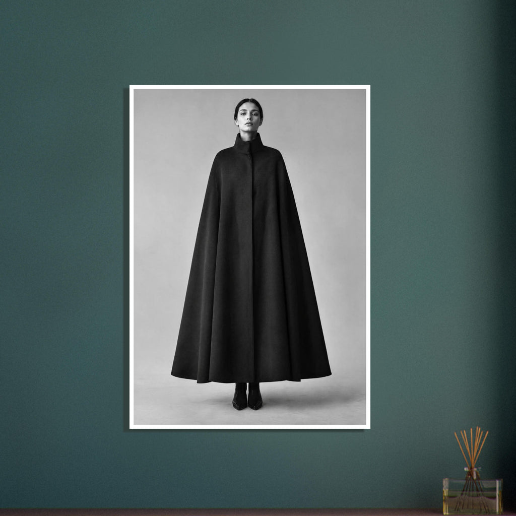 Woman wearing a long black cape with a high collar and black boots, standing against a plain grey studio backdrop.