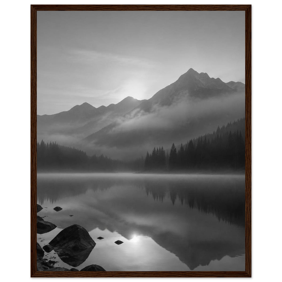 Mountain peaks and conifer forests reflected in a misty lake, with rocks in the foreground, in black and white.