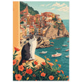 Grey and white cat on a flower-lined ledge above a teal harbour, small boats and colourful hillside buildings.