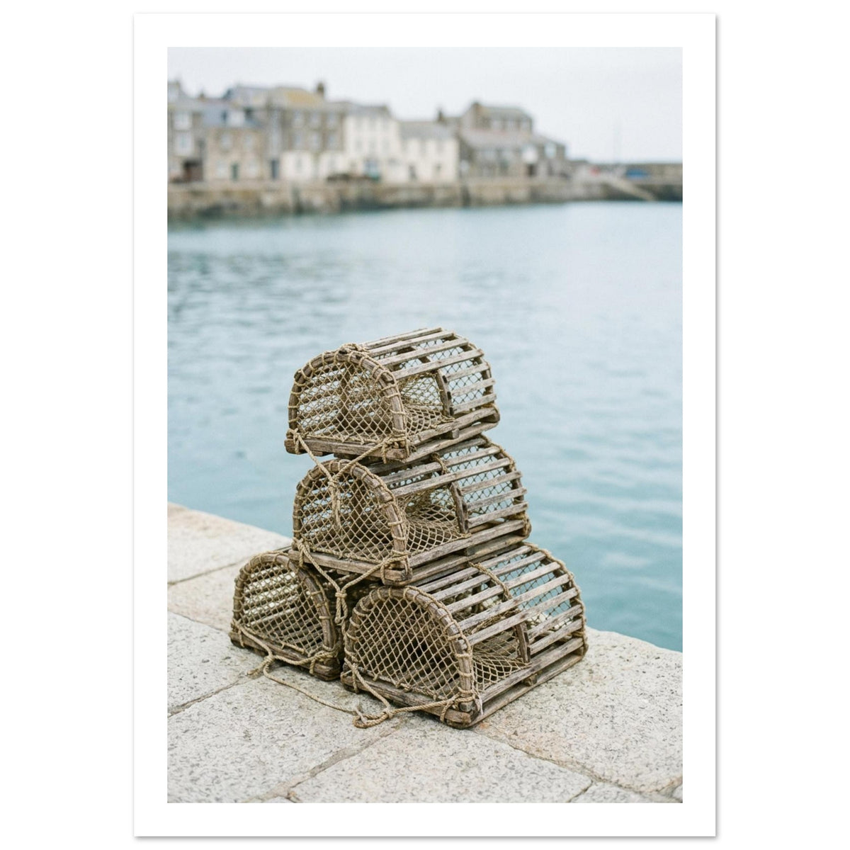 Four wooden lobster pots stacked on a stone quay beside pale blue harbour water, with blurred waterfront buildings beyond.