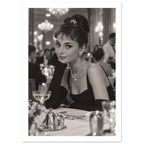 Woman in a black dress, long gloves and jewellery seated at a dining table, with blurred chandeliers behind her.