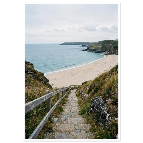 Stone steps with a wooden handrail descend through coastal grasses to a sandy beach, blue sea and rocky headlands.
