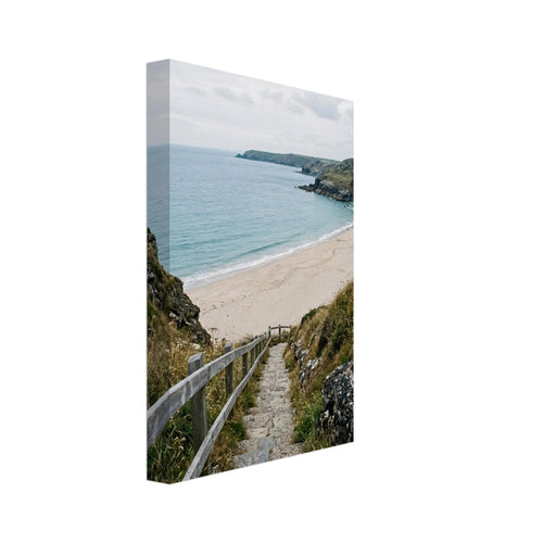 Stone steps with a wooden handrail descend through coastal grasses to a sandy beach, blue sea and rocky headlands.