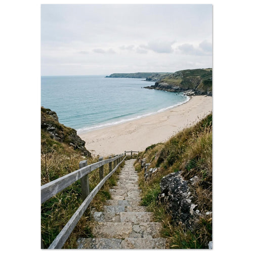 Stone steps with a wooden handrail descend through coastal grasses to a sandy beach, blue sea and rocky headlands.