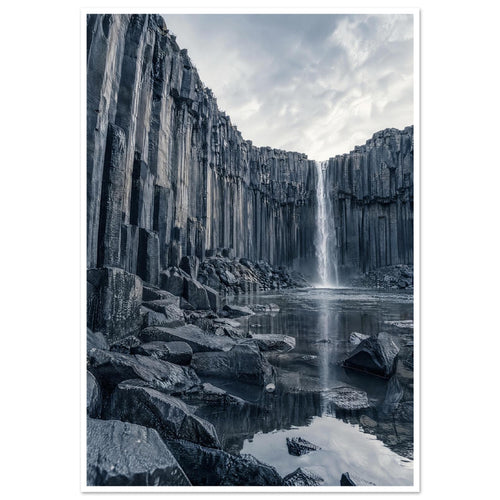 Waterfall cascading between tall basalt columns into a reflective pool, with dark foreground rocks and a cloudy sky.
