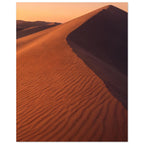 Desert sand dune with rippled orange slopes, a sweeping ridge and deep brown shadow beneath a peach sky.