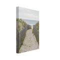 Sandy path with footprints between wooden fences and grassy dunes, leading towards a pale blue sea beneath a cloudy sky.