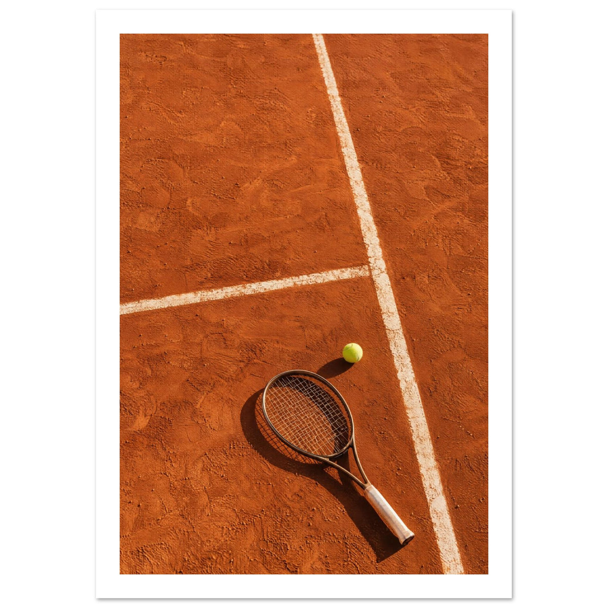 Tennis racket and yellow-green ball on an orange clay court, with white lines, footprints and strong shadows.
