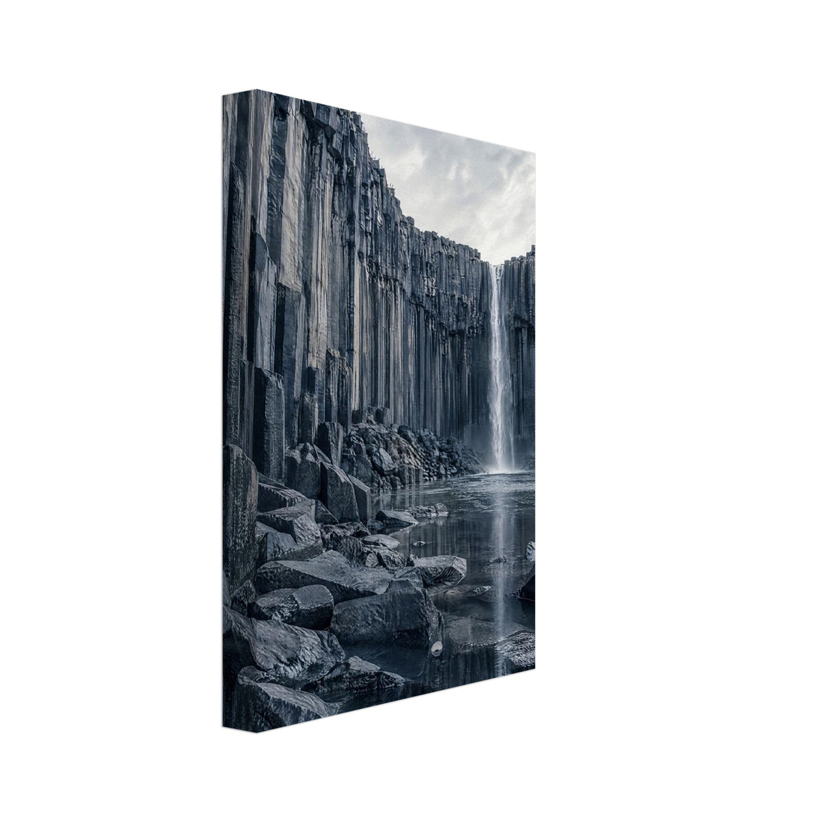 Waterfall cascading between tall basalt columns into a reflective pool, with dark foreground rocks and a cloudy sky.