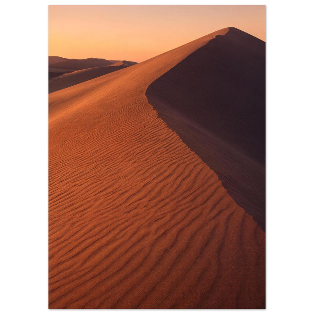 Desert sand dune with rippled orange slopes, a sweeping ridge and deep brown shadow beneath a peach sky.