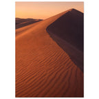 Desert sand dune with rippled orange slopes, a sweeping ridge and deep brown shadow beneath a peach sky.
