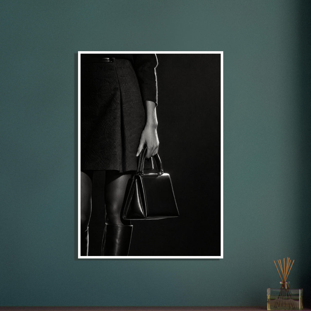 Cropped black and white photograph of a figure in a dark skirt and boots holding a glossy structured handbag.