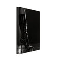Black and white photograph of a leg in a glossy knee-high stiletto boot on a reflective floor against a dark backdrop.
