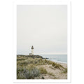 Lighthouse on a rocky headland beyond grassy sand dunes, with gentle waves and a wide, pale grey sky.