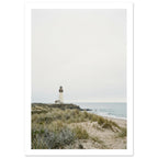 Lighthouse on a rocky headland beyond grassy sand dunes, with gentle waves and a wide, pale grey sky.