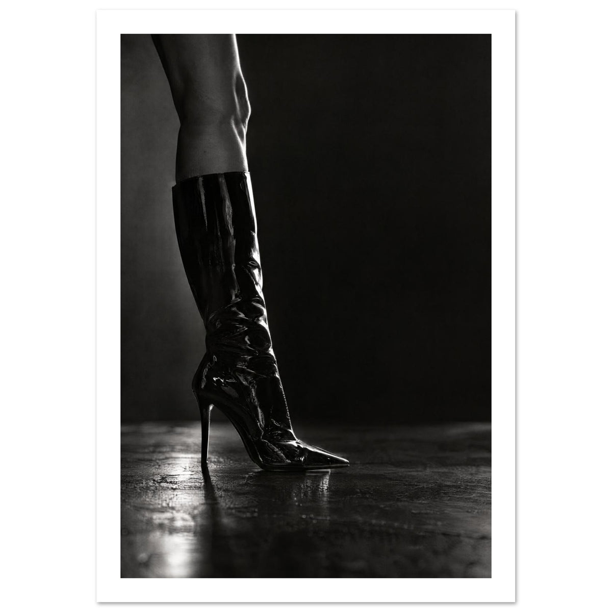 Black and white photograph of a leg in a glossy knee-high stiletto boot on a reflective floor against a dark backdrop.