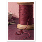 Wooden spool of burgundy thread with a loose looping strand, scissors and a bowl of pins against a pink background.