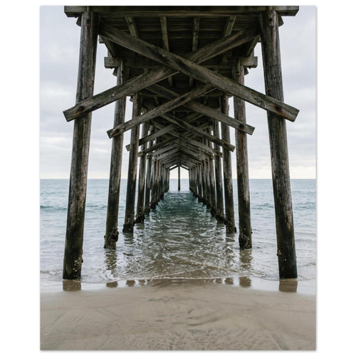 Timber pier viewed from beneath, with crossed wooden beams receding over gentle sea waves and wet sand under a grey sky.