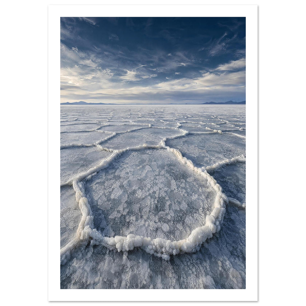 Salt flats with raised polygon-shaped ridges stretching towards distant mountains beneath a blue, cloud-streaked sky.