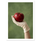 Red apple resting in a cream-gloved hand, with a bare forearm below and a softly blurred green background.