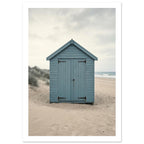 Blue wooden beach hut with closed doors on pale sand, with dune grasses, sea waves and an overcast sky.