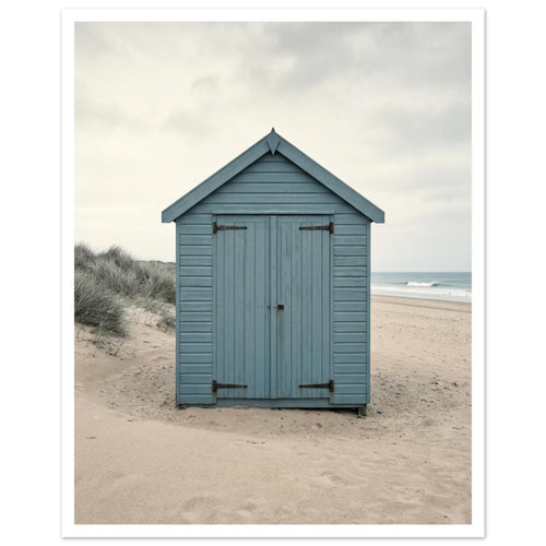 Blue wooden beach hut with closed doors on pale sand, with dune grasses, sea waves and an overcast sky.