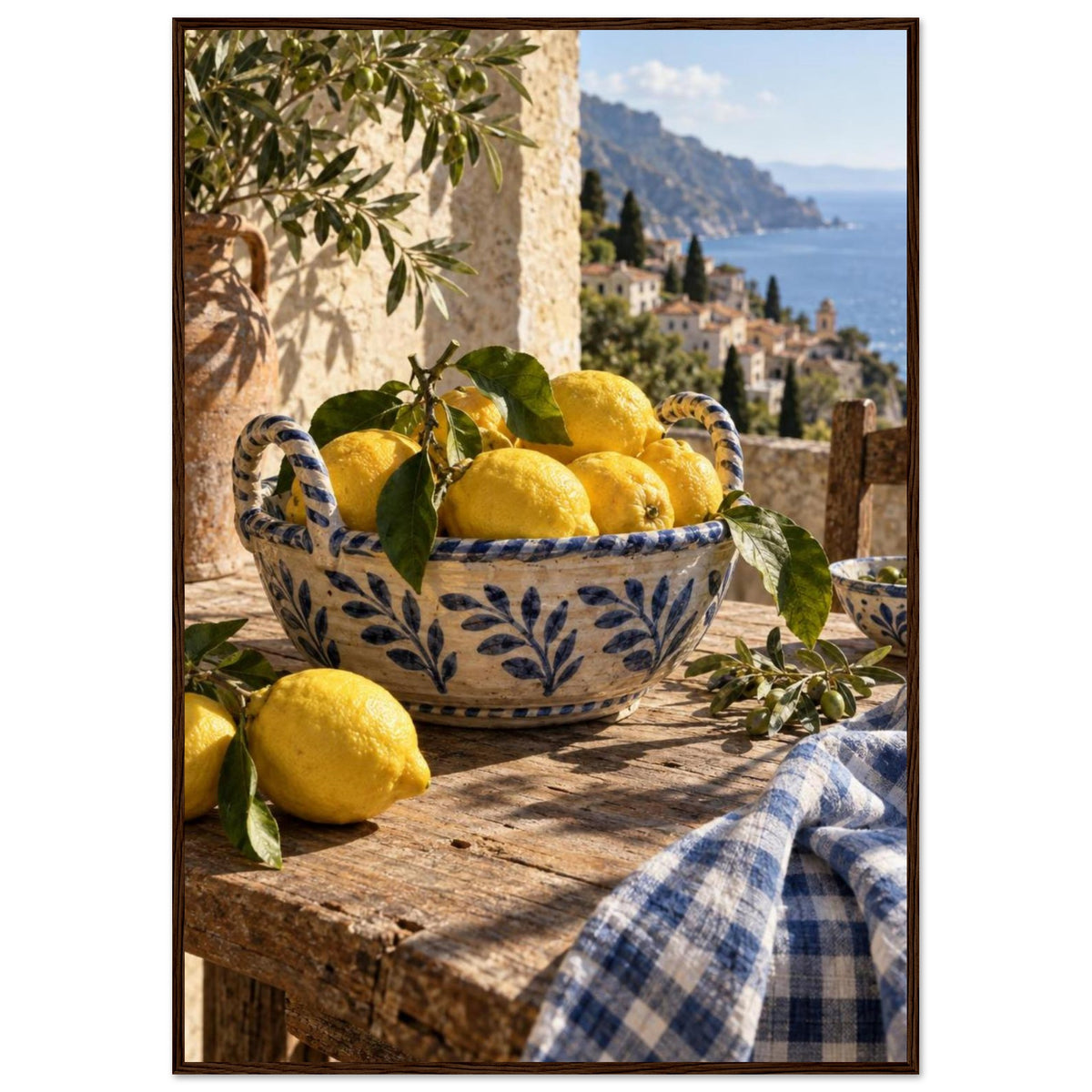 Lemons in a blue-and-white bowl on a wooden table, with olive branches, checked cloth and a coastal village beyond.
