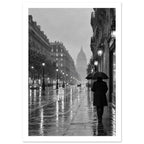 Umbrella-carrying figure on a rainy boulevard with ornate buildings, glowing lamps and wet street reflections in black and wh