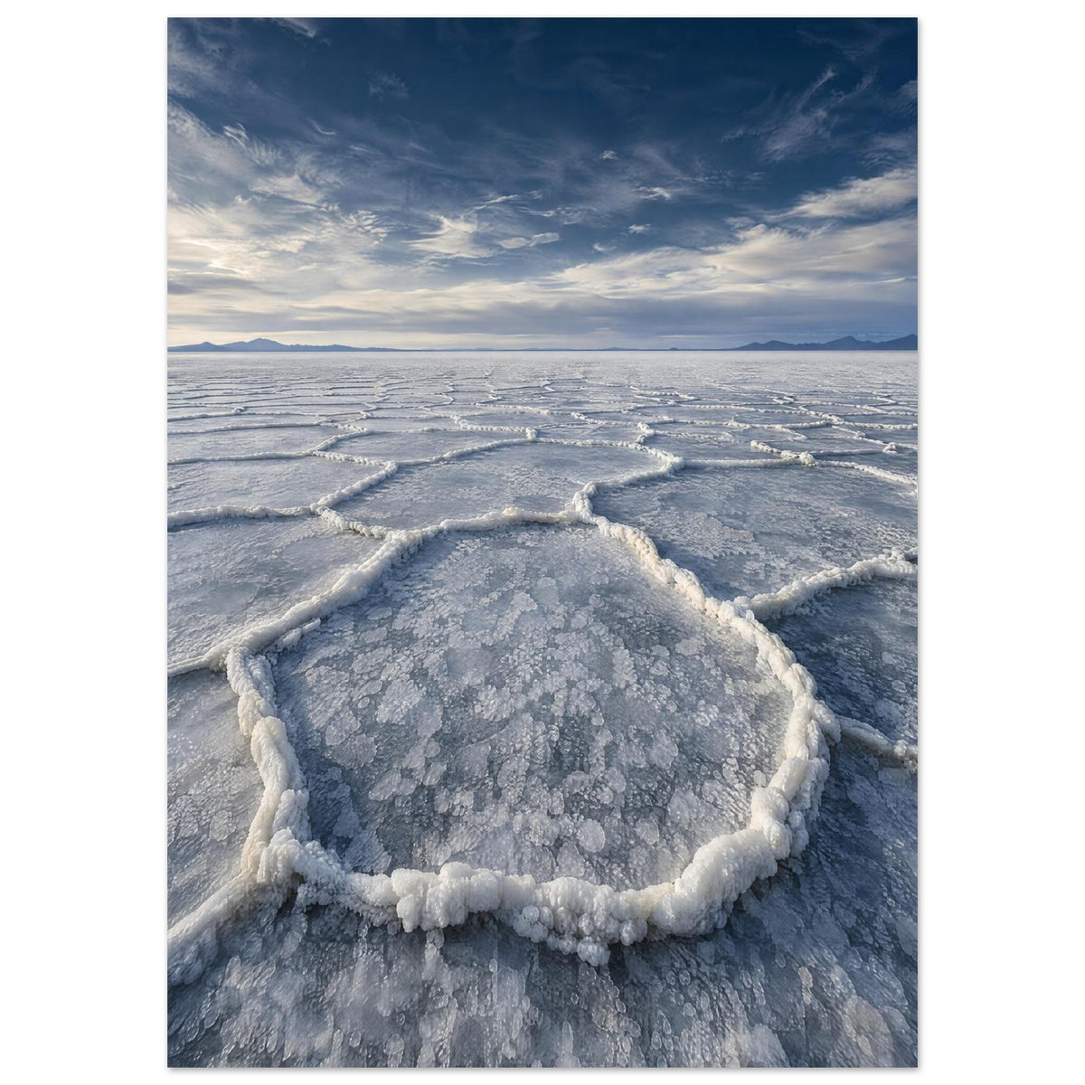 Salt flats with raised polygon-shaped ridges stretching towards distant mountains beneath a blue, cloud-streaked sky.