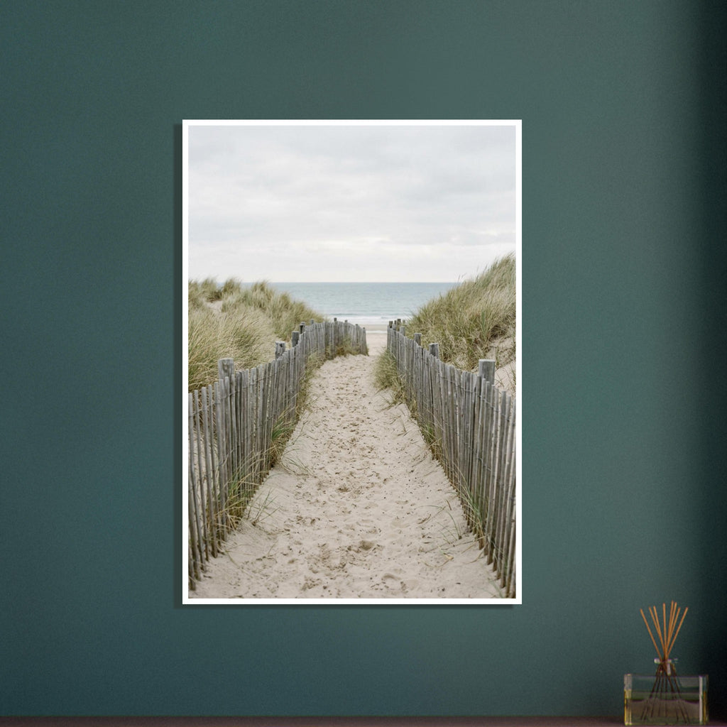 Sandy path with footprints between wooden fences and grassy dunes, leading towards a pale blue sea beneath a cloudy sky.
